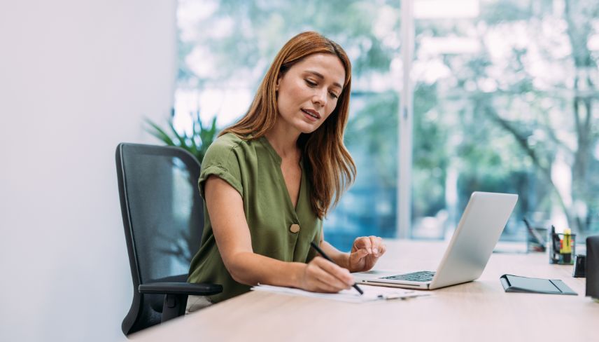 A woman sitting at a desk, writing in a notebook while using a laptop in a bright office setting.